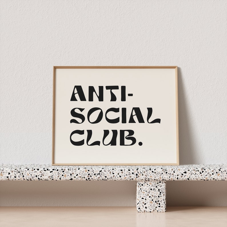 May include: A framed print with the text "ANTI-SOCIAL CLUB." in black stylized lettering on a white background. The frame is a light wood color and the print is sitting on a terrazzo shelf.