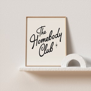 May include: A framed print with a white background and black text that reads "The Homebody Club" with a small star symbol. The print is sitting on a white shelf with a white decorative arch in front of it.