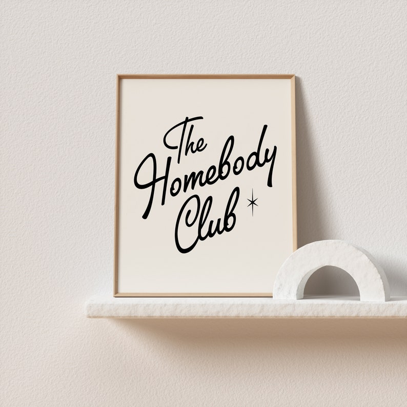 May include: A framed print with a white background and black text that reads "The Homebody Club" with a small star symbol. The print is sitting on a white shelf with a white decorative arch in front of it.