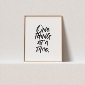 May include: A framed print with a white background and black calligraphy that reads "One thing at a time."