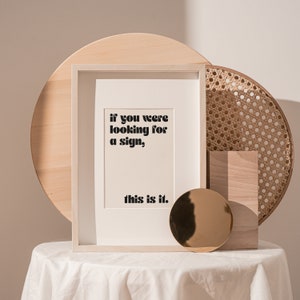 May include: A white framed print with the text "if you were looking for a sign, this is it." The print is sitting on a white table with a wooden tray and a woven basket behind it.