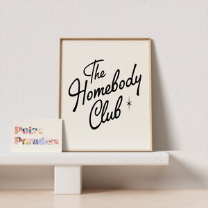 May include: A framed print with a white background and black text that reads "The Homebody Club" with a small star next to the word "Club". A small card with the text "Poire Paradise" is sitting on a white shelf in front of the print.
