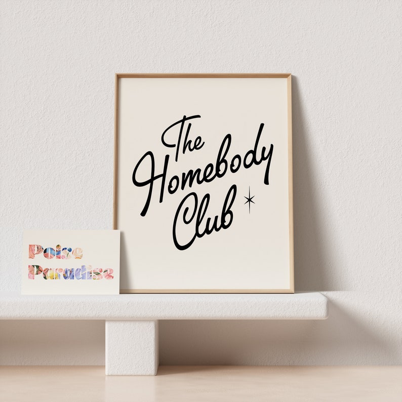 May include: A framed print with a white background and black text that reads "The Homebody Club" with a small star next to the word "Club". A small card with the text "Poire Paradise" is sitting on a white shelf in front of the print.