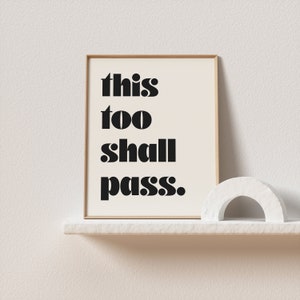 K&ouml;nnte beinhalten: Ein gerahmter Druck mit dem Text "this too shall pass." in schwarzen Fettdruck auf wei&szlig;em Hintergrund. Der Druck wird auf einem wei&szlig;en Regal mit einem wei&szlig;en Dekorationsobjekt pr&auml;sentiert.