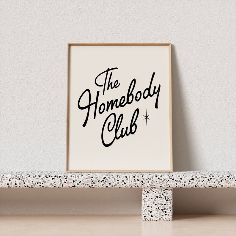 May include: A framed print with a white background and black text that reads "The Homebody Club" with a small star next to the word "Club".
