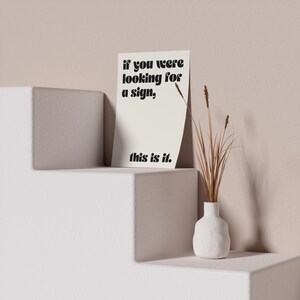 May include: A white card with black text that reads "if you were looking for a sign, this is it." The card is sitting on a white shelf with a white vase with dried brown grass on the right side.
