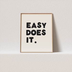 以下が含まれることがあります： 白い背景に黒い太字で「EASY DOES IT.」と書かれた額装されたプリント