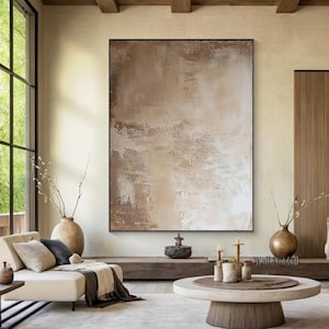 May include: Large abstract painting in shades of brown and beige, framed in a black border. The artwork hangs in a modern living room with a white sofa, wooden accents, and a round coffee table.