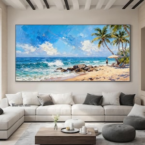 May include: Large oil painting of a beach scene with a bright blue sky and white clouds. The ocean waves crash against the shore, and palm trees are visible on the beach. The painting is framed and hangs above a white sofa.