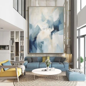 May include: Abstract painting with blue and white hues. The painting is framed and hanging on a wall in a modern living room. The room features a blue sofa, a coffee table, and a yellow armchair.
