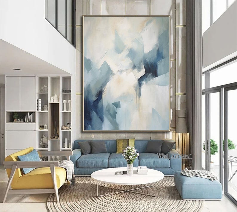 May include: Abstract painting with blue and white hues. The painting is framed and hanging on a wall in a modern living room. The room features a blue sofa, a coffee table, and a yellow armchair.