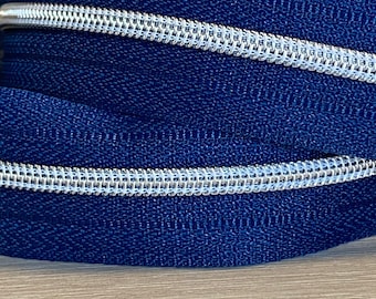 May include: A close-up of a blue zipper with a silver zipper pull. The zipper tape is a dark blue color and the zipper teeth are silver.