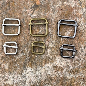 May include: An assortment of six metal slider buckles in various finishes: silver, antique brass, and black. The buckles are rectangular with a single bar, suitable for straps or belts. The buckles are displayed on a textured, brown stone surface.