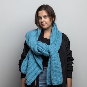 Könnte beinhalten: Eine Frau trägt eine schwarze Jacke mit einem blauen Strick Schal um den Hals. Der Schal ist hellblau und hat eine strukturierte Strickart.