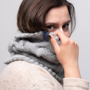 以下が含まれることがあります： グレーのニットのタートルネックのストールを着用した女性。ストールには「Knitwear」と書かれた青いラベルが付いています。ベージュのセーターを着ており、片手で顔を隠しています。