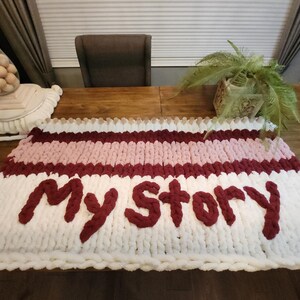 Peut inclure: Une couverture en tricot &eacute;pais avec les mots "My Story" en fil bordeaux. La couverture pr&eacute;sente des rayures blanches, rose clair et bordeaux. La couverture est pos&eacute;e sur une table en bois.