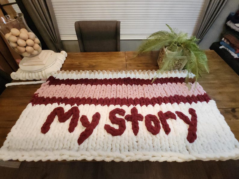 Peut inclure: Une couverture en tricot &eacute;pais avec les mots "My Story" en fil bordeaux. La couverture pr&eacute;sente des rayures blanches, rose clair et bordeaux. La couverture est pos&eacute;e sur une table en bois.