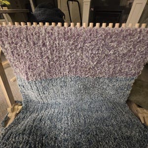 Peut inclure: Une couverture moelleuse faite &agrave; la main, dans des tons de lavande, de bleu et de gris, fabriqu&eacute;e sur un m&eacute;tier &agrave; tisser en bois. La laine textur&eacute;e cr&eacute;e un aspect doux et invitant. La couverture est partiellement finie, montrant le processus de tissage.