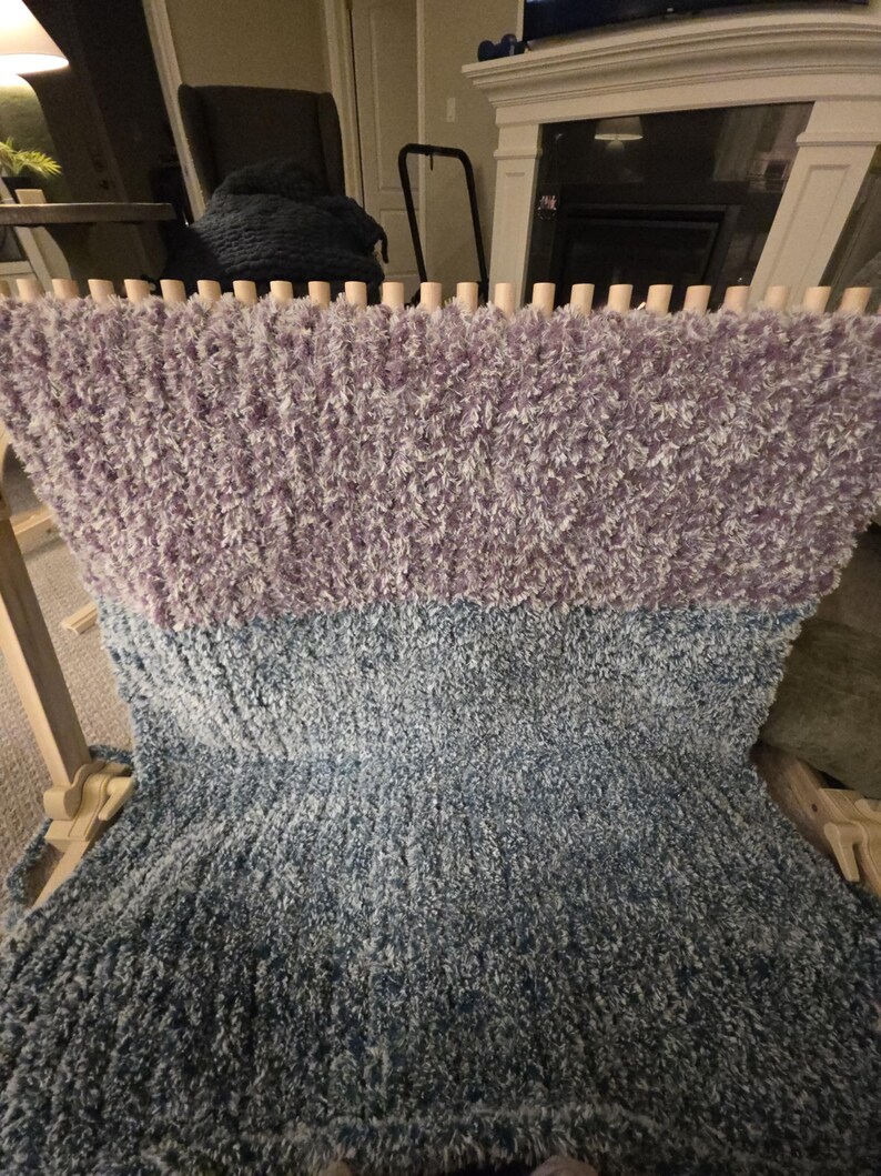 Peut inclure: Une couverture moelleuse faite &agrave; la main, dans des tons de lavande, de bleu et de gris, fabriqu&eacute;e sur un m&eacute;tier &agrave; tisser en bois. La laine textur&eacute;e cr&eacute;e un aspect doux et invitant. La couverture est partiellement finie, montrant le processus de tissage.