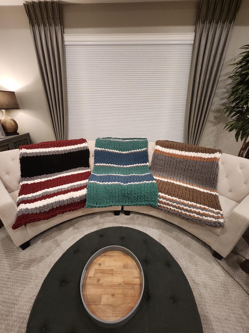 Peut inclure: Trois couvertures en tricot &eacute;pais de couleurs vari&eacute;es sont pos&eacute;es sur un canap&eacute; blanc. L'une pr&eacute;sente des rayures bordeaux, blanches et grises. Une autre a des rayures vertes et bleues. La troisi&egrave;me a des rayures marron, grises et blanches.