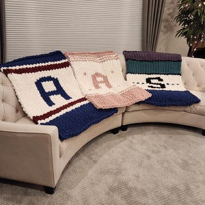 Puede incluir: Tres mantas de punto con las letras "A A S" deletreadas en hilo blanco sobre un sofá de color crema. Las mantas son azul marino, rosa y turquesa.