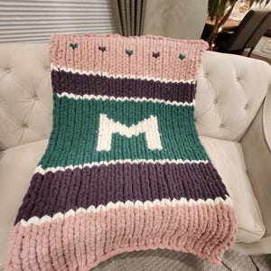 Peut inclure: Une couverture en tricot &eacute;pais aux tons rose, vert, violet et blanc, avec un grand "M" blanc au centre. La couverture, &agrave; motifs ray&eacute;s, est pos&eacute;e sur un canap&eacute; cr&egrave;me. De petits c&oelig;urs verts ornent le haut.