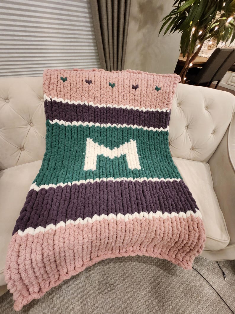 Peut inclure: Une couverture en tricot &eacute;pais aux tons rose, vert, violet et blanc, avec un grand "M" blanc au centre. La couverture, &agrave; motifs ray&eacute;s, est pos&eacute;e sur un canap&eacute; cr&egrave;me. De petits c&oelig;urs verts ornent le haut.