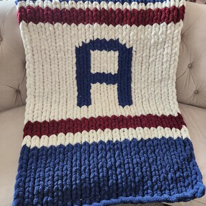 Peut inclure: Une couverture en tricot &eacute;pais avec des rayures bleues, blanches et bordeaux, avec un grand "A" bleu au centre. La couverture est faite de fil &eacute;pais et doux. Le sch&eacute;ma de couleurs rappelle une &eacute;quipe sportive.
