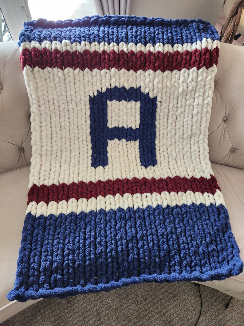Peut inclure: Une couverture en tricot &eacute;pais avec des rayures bleues, blanches et bordeaux, avec un grand "A" bleu au centre. La couverture est faite de fil &eacute;pais et doux. Le sch&eacute;ma de couleurs rappelle une &eacute;quipe sportive.