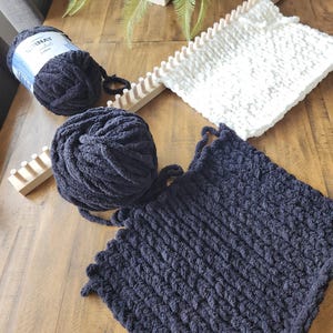 May include: A still-life image featuring knitting supplies. A dark navy blue knitted square and a ball of matching yarn are displayed alongside a white knitted square and a wooden knitting loom. The yarn label is visible.