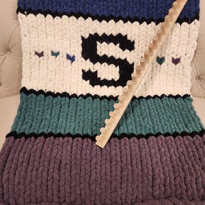 Peut inclure: Une couverture en tricot &eacute;pais dans les tons de bleu, blanc, sarcelle et violet, avec une grande lettre "S" noire et de petits c&oelig;urs. Un m&eacute;tier &agrave; tricoter en bois repose sur la couverture. La couverture est en fil &eacute;pais et doux.