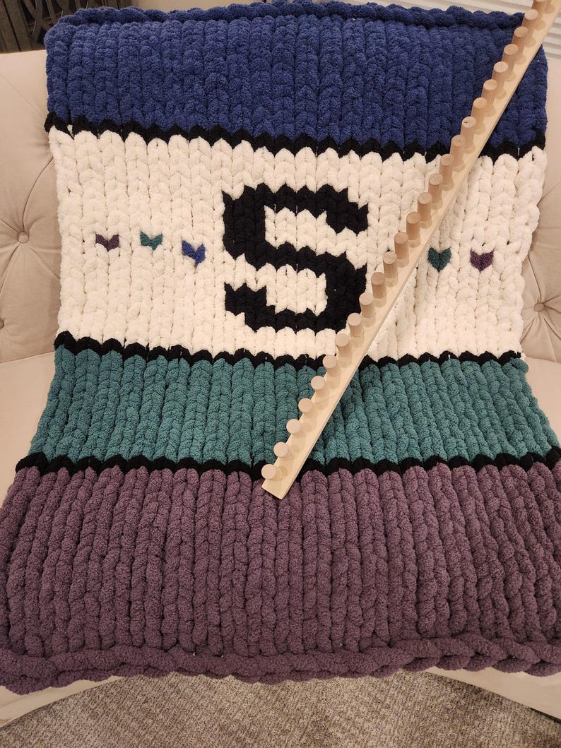 Peut inclure: Une couverture en tricot &eacute;pais dans les tons de bleu, blanc, sarcelle et violet, avec une grande lettre "S" noire et de petits c&oelig;urs. Un m&eacute;tier &agrave; tricoter en bois repose sur la couverture. La couverture est en fil &eacute;pais et doux.