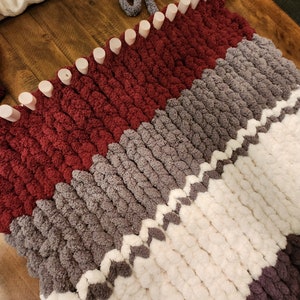 Peut inclure: Gros plan sur une couverture tricot&eacute;e &agrave; la main en cours de r&eacute;alisation, avec un motif en blocs de couleurs. La couverture est faite avec de la laine &eacute;paisse et &eacute;paisse dans les tons bordeaux, gris, blanc et violet. Le m&eacute;tier &agrave; tricoter avec des chevilles en bois est visible.