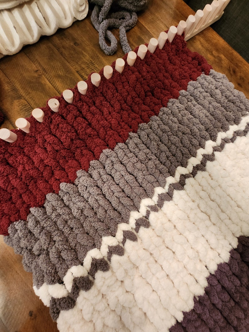 Peut inclure: Gros plan sur une couverture tricot&eacute;e &agrave; la main en cours de r&eacute;alisation, avec un motif en blocs de couleurs. La couverture est faite avec de la laine &eacute;paisse et &eacute;paisse dans les tons bordeaux, gris, blanc et violet. Le m&eacute;tier &agrave; tricoter avec des chevilles en bois est visible.
