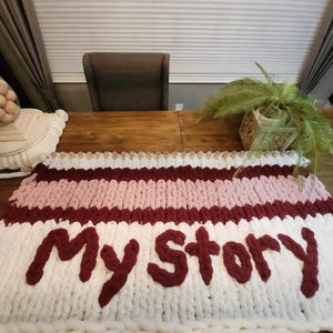 Peut inclure: Une couverture en tricot &eacute;pais avec des rayures blanches, roses et bordeaux, avec les mots "My Story" en fil bordeaux. La couverture est pos&eacute;e sur une table en bois.