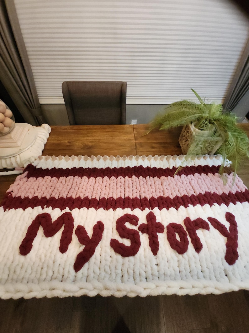 Peut inclure: Une couverture en tricot &eacute;pais avec des rayures blanches, roses et bordeaux, avec les mots "My Story" en fil bordeaux. La couverture est pos&eacute;e sur une table en bois.