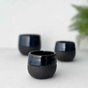 Könnte beinhalten: Drei kleine, abgerundete Keramikbecher mit zweifarbigem Design. Der obere Teil jedes Bechers ist mit einer glatten, glänzenden schwarzen Glasur versehen, während der untere Teil eine matte, texturierte schwarze Oberfläche aufweist.