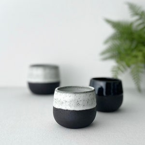 Könnte beinhalten: Drei kleine, abgerundete Keramikbecher in zweifarbigem Design. Jeder Becher hat eine mattschwarze Basis und einen gesprenkelten, cremeweißen glasierten oberen Teil. Die Becher haben eine einfache, henkellose, schalenartige Form.