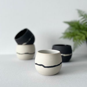 Pode incluir: Três pequenas chávenas de cerâmica com formato arredondado. Uma chávena é branca com uma linha preta horizontal, outra é preta com uma linha branca, e a terceira é uma chávena branca empilhada sob uma preta, ambas com linhas horizontais contrastantes.