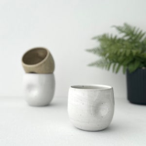 Könnte beinhalten: Drei Keramikbecher im minimalistischen Stil. Ein weißer Becher steht im Fokus, mit einer strukturierten Oberfläche und einer abgerundeten Form. Zwei weitere Becher befinden sich im Hintergrund, einer weiß und einer beige, übereinander gestapelt. Ein Farn ist im Hintergrund.