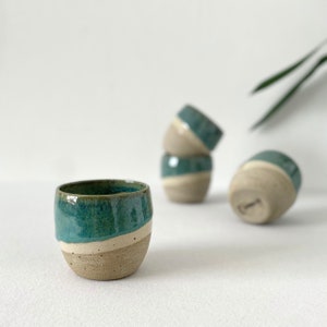 Op de afbeelding: Vier keramische bekers met een turquoise glazuur en een witte band rond de bovenkant. De bekers staan op een wit oppervlak.