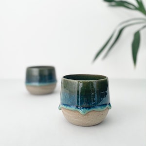 Pode incluir: Duas canecas de cerâmica com um esmalte verde e azul. As canecas têm uma forma arredondada e um lábio ligeiramente alargado. O esmalte tem um efeito de gotejamento ao redor da borda das canecas.
