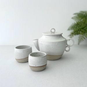 May include: A white ceramic tea set comprising a teapot and two cups. The teapot features a rounded body, a lid with a ring handle, and a curved spout. The cups and teapot have a light brown base. The set is designed for serving tea.