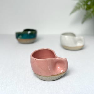 Pode incluir: Um pote de cerâmica rosa com base cinza está em foco, com dois outros potes de cerâmica no fundo. Um é branco e o outro é turquesa e verde. Os potes têm um formato único e orgânico.