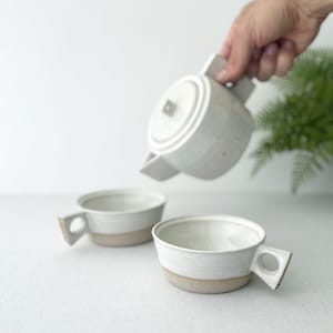 May include: White ceramic teapot being poured into two white ceramic mugs with handles. The teapot and mugs have a speckled glaze.