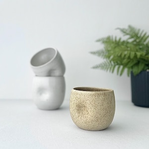 May include: A speckled beige ceramic cup with a rounded shape. The cup is sitting on a white surface.
