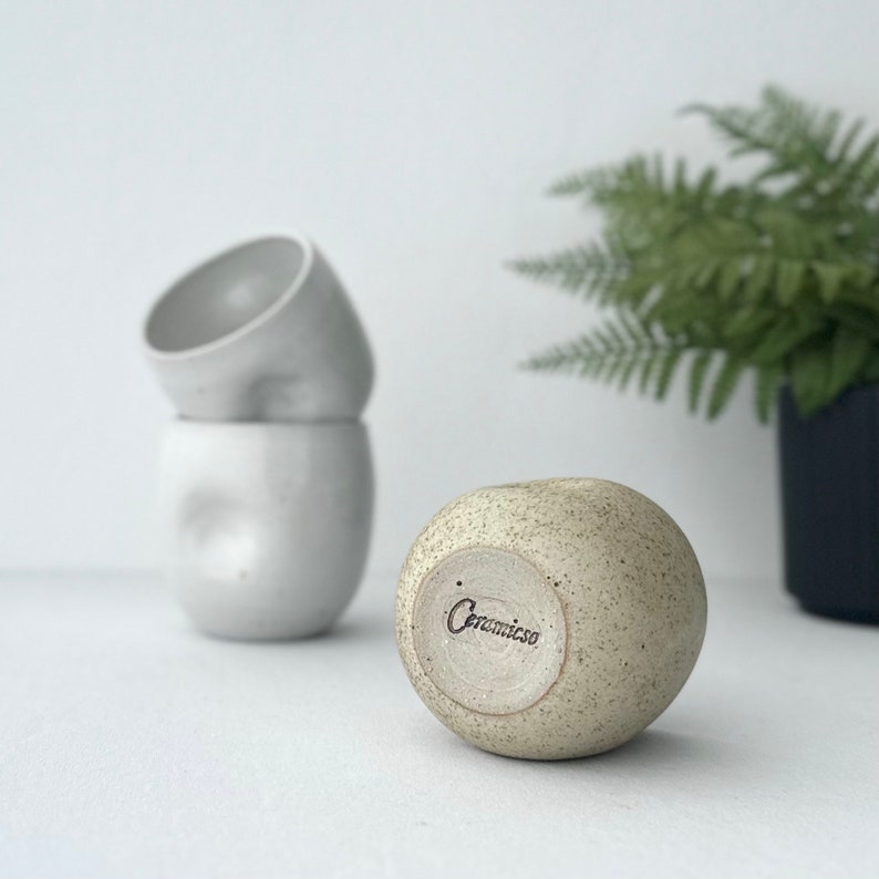 May include: A speckled beige ceramic cup with the word "Ceramico" stamped on the bottom. The cup is sitting on a white surface next to two white ceramic cups.
