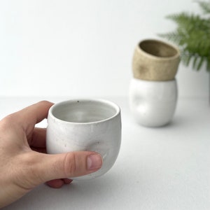 May include: A white ceramic cup with a slightly textured surface. The cup is being held in a hand. There are two other white ceramic cups in the background.