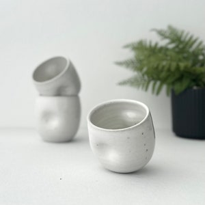May include: Three white ceramic mugs with a textured, uneven surface. The mugs are stacked on top of each other, with the top mug slightly tilted.
