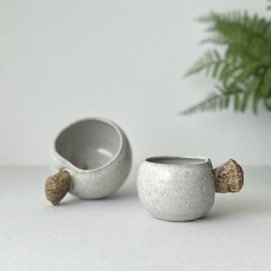 Puede incluir: Dos tazas de cerámica blanca con un acabado moteado y un asa de madera marrón. Las tazas están sobre una superficie blanca.